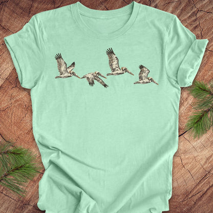 A mint green short-sleeve t-shirt with a graphic print of five flying pelicans, laid flat on a rustic wood background with pine needles in the corners.