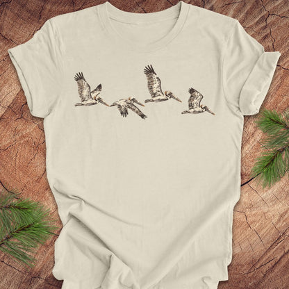 An off-white short-sleeve t-shirt featuring a graphic print of five flying birds in black and brown, laid flat on a rustic wood background with pine needles in the corners.