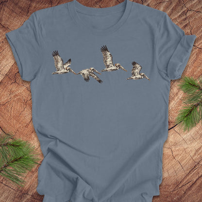 A slate blue short-sleeve t-shirt featuring a graphic print of five flying pelicans, laid flat on a rustic wood background with pine branches in the corners.