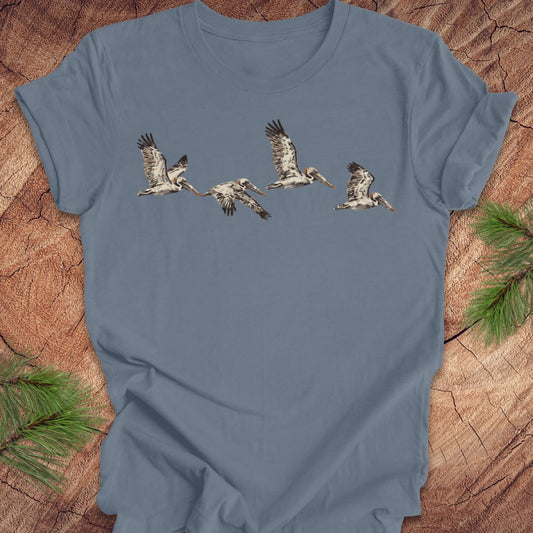 A slate blue short-sleeve t-shirt featuring a graphic print of five flying pelicans, laid flat on a rustic wood background with pine branches in the corners.