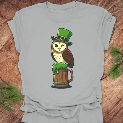 Gray t-shirt with an owl wearing a green leprechaun hat and holding a beer mug on a wooden surface.