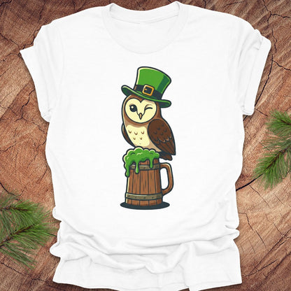 White t-shirt with an owl design wearing a green leprechaun hat and standing on a wooden barrel of green beer, on a wooden surface.