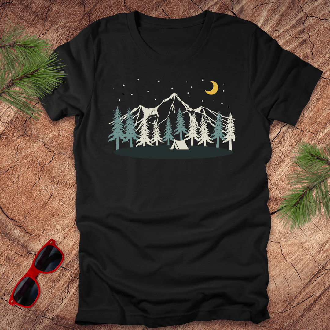 Black t-shirt with mountain and tent design on a wooden surface with sunglasses and pine branches.
