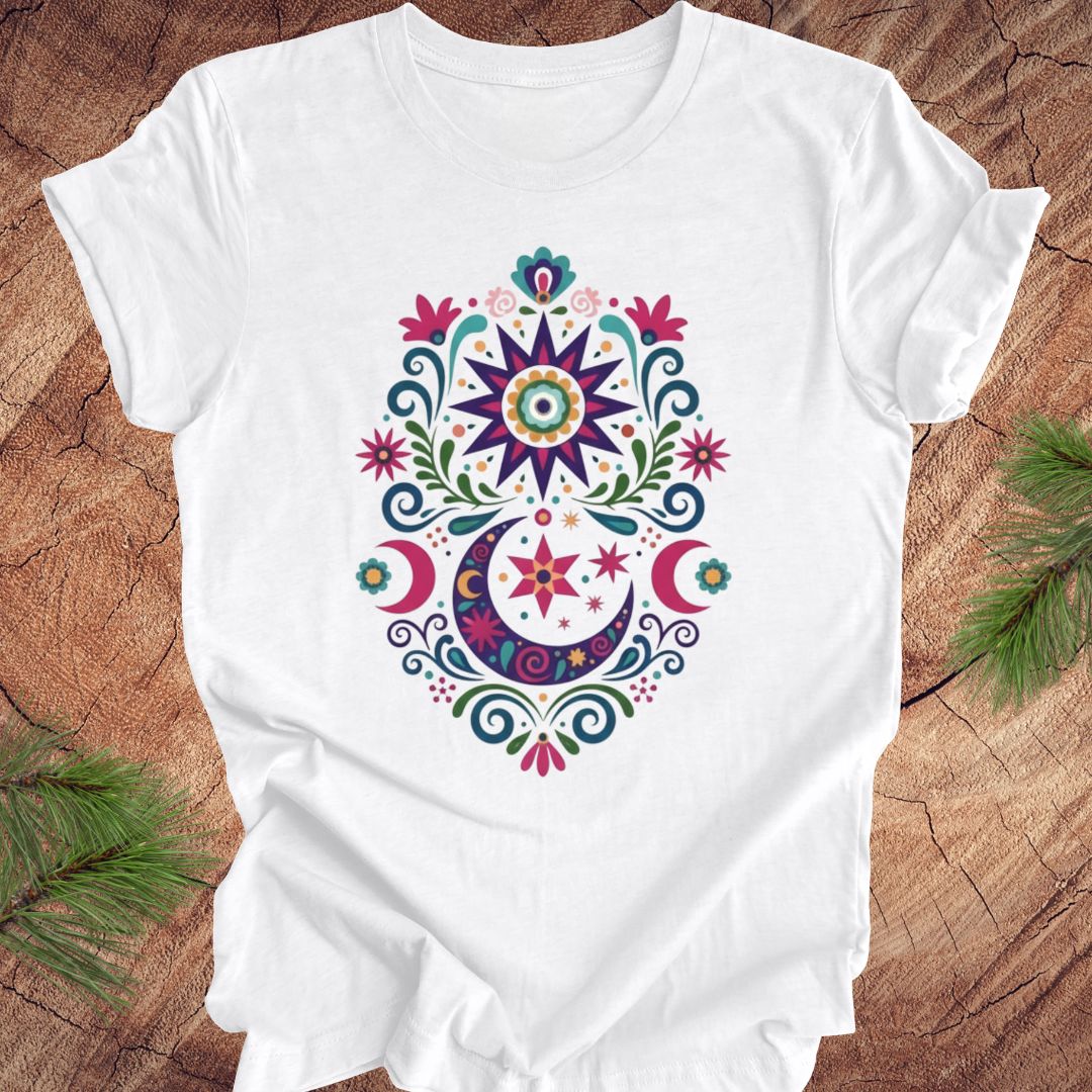 White t-shirt with a colorful mandala design on a wooden surface with green leaves.