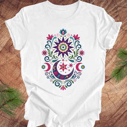 White t-shirt with a colorful mandala design on a wooden surface with green leaves.