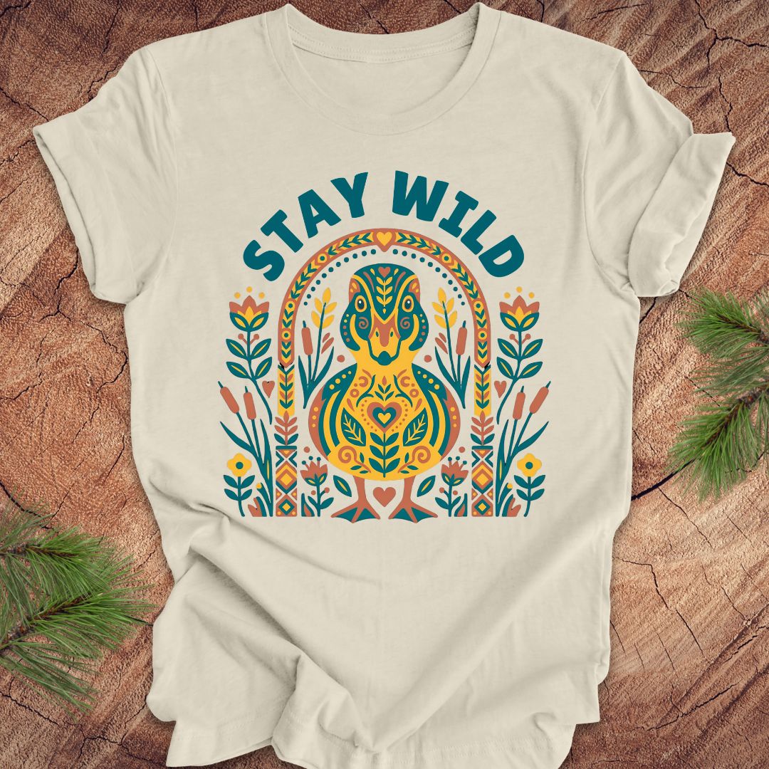 Beige t-shirt with colorful duck graphic and 'Stay Wild' text on a wooden surface.