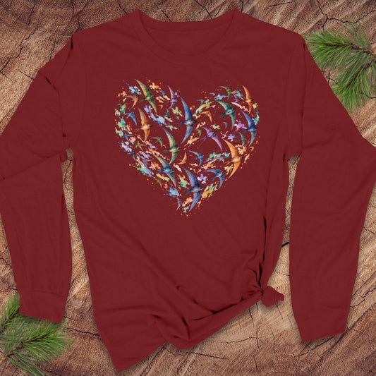 A long-sleeve crewneck t-shirt in burgundy red, featuring a large heart-shaped graphic on the chest made of colorful, overlapping bird silhouettes. The shirt is styled flat on a rustic wood background with pine needles in the corners.