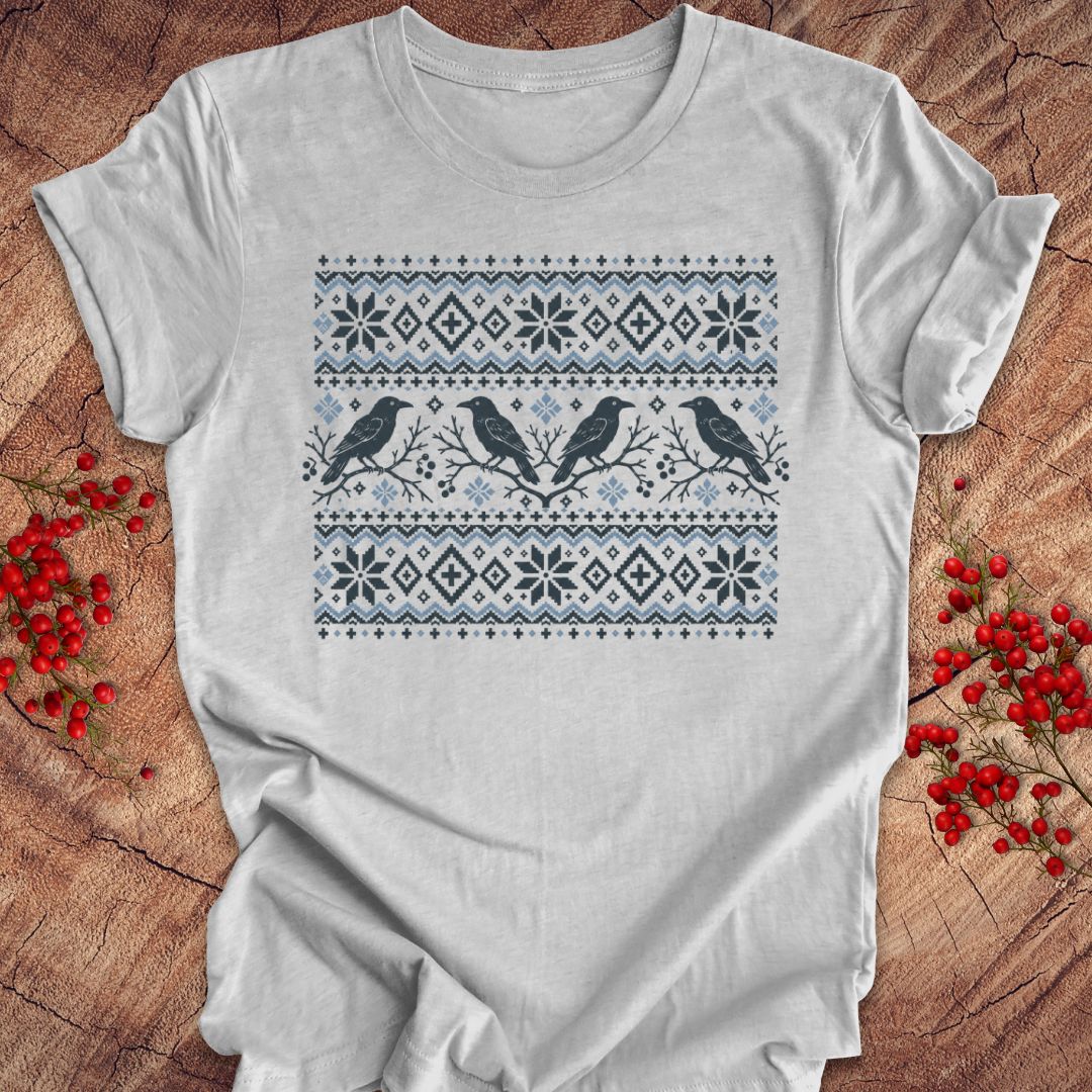 A light gray short-sleeve t-shirt with a Nordic-style holiday print featuring two black birds and snowflake patterns in navy and light blue, laid flat on a rustic wood background with red berries.