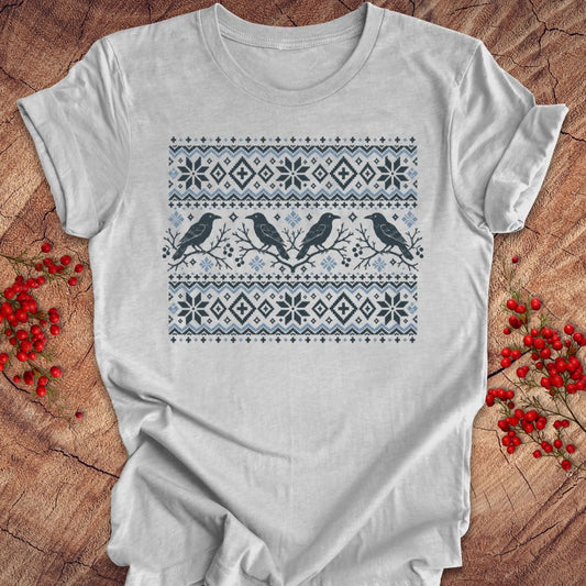 A light gray short-sleeve t-shirt with a Nordic-style holiday print featuring two black birds and snowflake patterns in navy and light blue, laid flat on a rustic wood background with red berries.