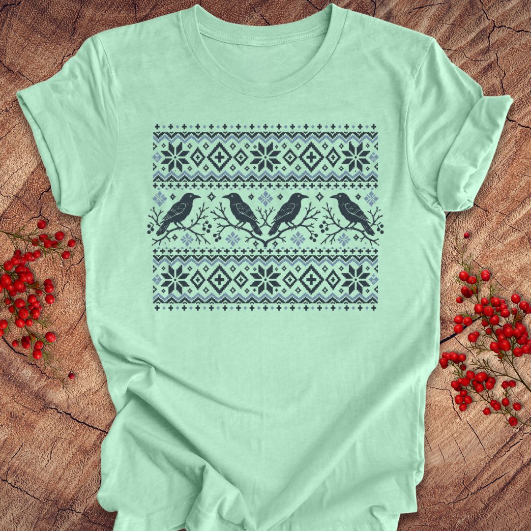 A mint green short-sleeve t-shirt with a dark gray and light blue Nordic-style pattern featuring birds and snowflakes, laid flat on a rustic wood background with red berries.