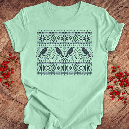 A mint green short-sleeve t-shirt with a dark gray and light blue Nordic-style pattern featuring birds and snowflakes, laid flat on a rustic wood background with red berries.