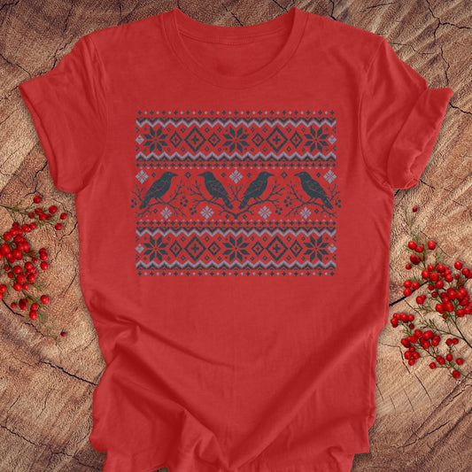 A red short-sleeve crewneck t-shirt with a festive Nordic-style knit pattern featuring two black birds and snowflakes, laid flat on a rustic wood background with red berries.