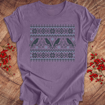 A lavender purple short-sleeve t-shirt with a winter-themed graphic print featuring three birds and snowflake patterns, laid flat on a rustic wood background with red berries.