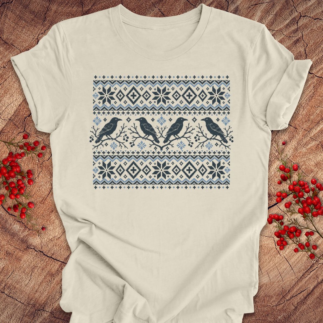 An off-white short-sleeve t-shirt with a blue and navy Nordic-style pattern featuring snowflakes and birds, laid flat on a rustic wood background with red berries.