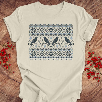 An off-white short-sleeve t-shirt with a blue and navy Nordic-style pattern featuring snowflakes and birds, laid flat on a rustic wood background with red berries.
