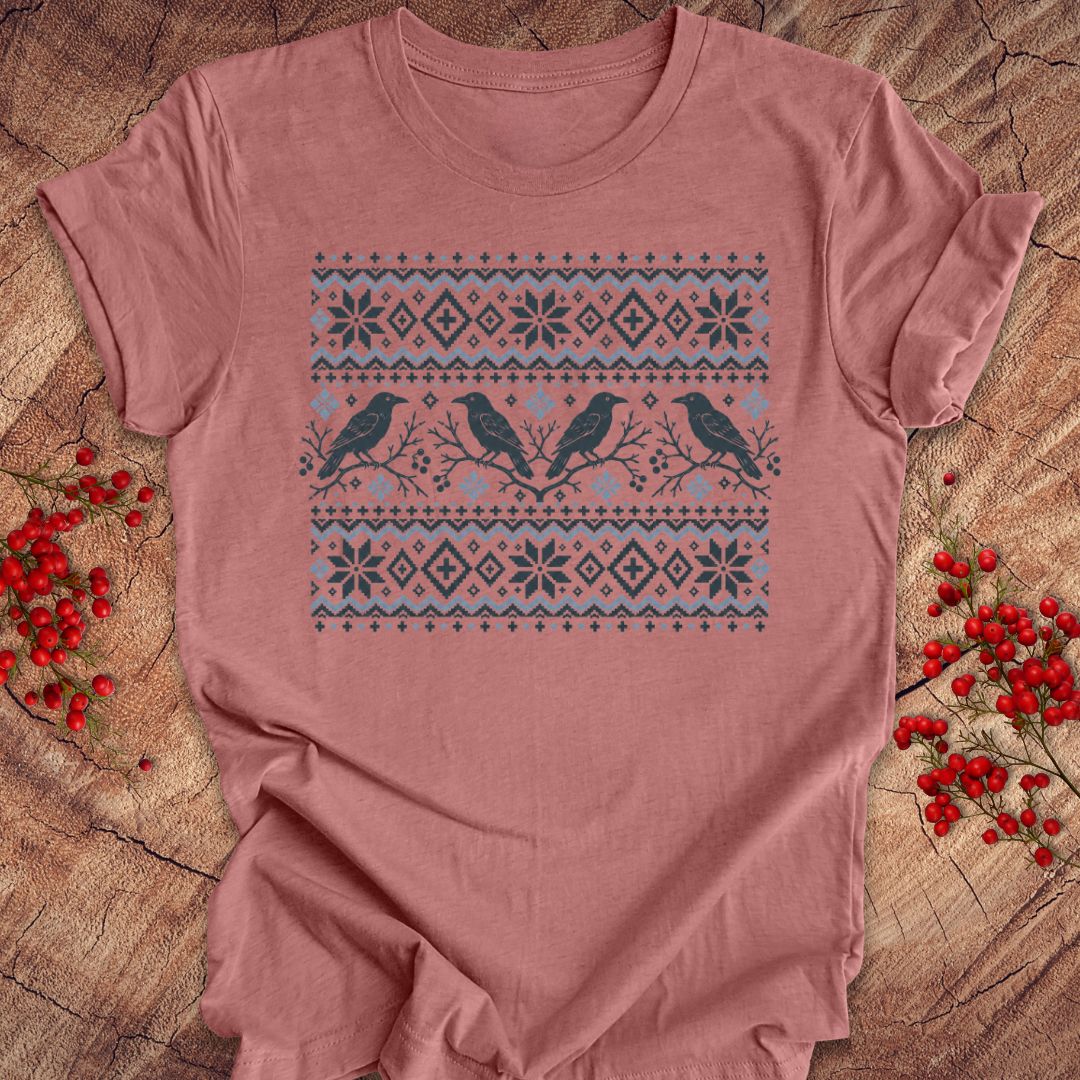 A dusty rose short-sleeve t-shirt with a winter-themed bird graphic, laid flat on a rustic wood background with red berries.