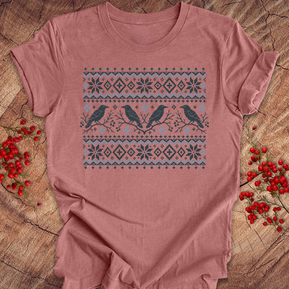 A dusty rose short-sleeve t-shirt with a winter-themed bird graphic, laid flat on a rustic wood background with red berries.