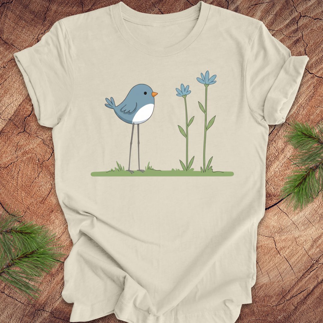 Beige t-shirt with a blue bird and flowers graphic on a wooden surface.