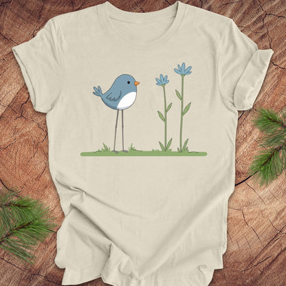 Beige t-shirt with a blue bird and flowers graphic on a wooden surface.