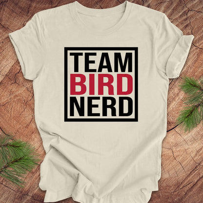 Beige t-shirt with 'Team Bird Nerd' text on a wooden surface with greenery.