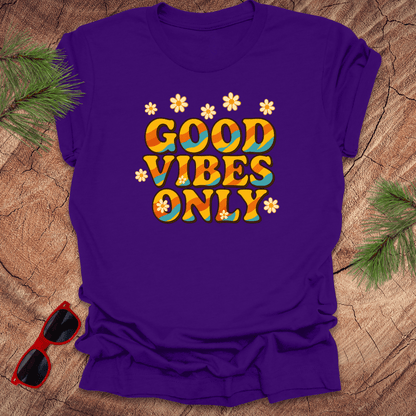 Purple t-shirt with 'Good Vibes Only' text on a wooden surface with sunglasses and greenery.