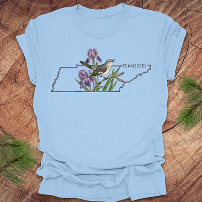 Baby blue t-shirt with a graphic of Tennessee's state shape featuring a mockingbird and iris flowers on a wooden surface.