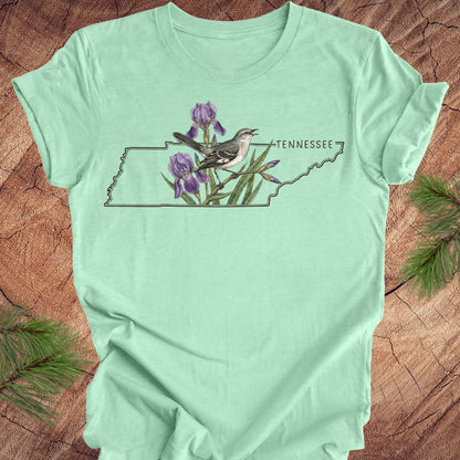 Mint green t-shirt with a graphic of a bird and flowers on a map of Tennessee, placed on a wooden surface.