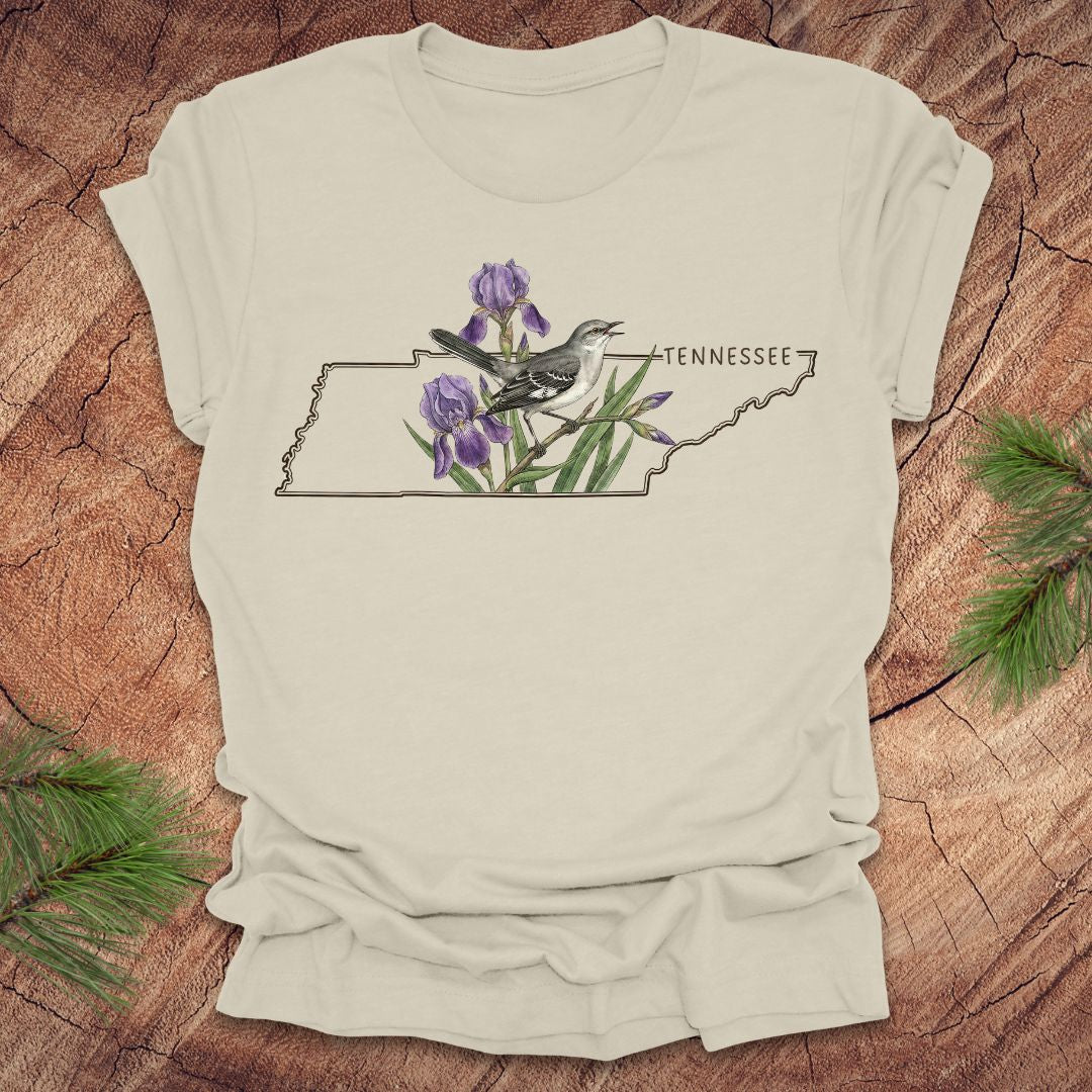 Beige t-shirt with a graphic of Tennessee's state shape featuring a mockingbird and iris flowers on a wooden surface.
