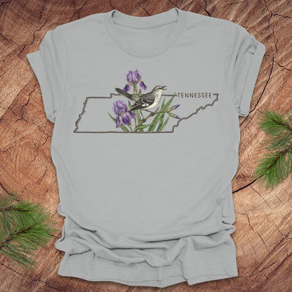 Silver t-shirt with a graphic of Tennessee's state shape featuring a mockingbird and iris flowers on a wooden surface.