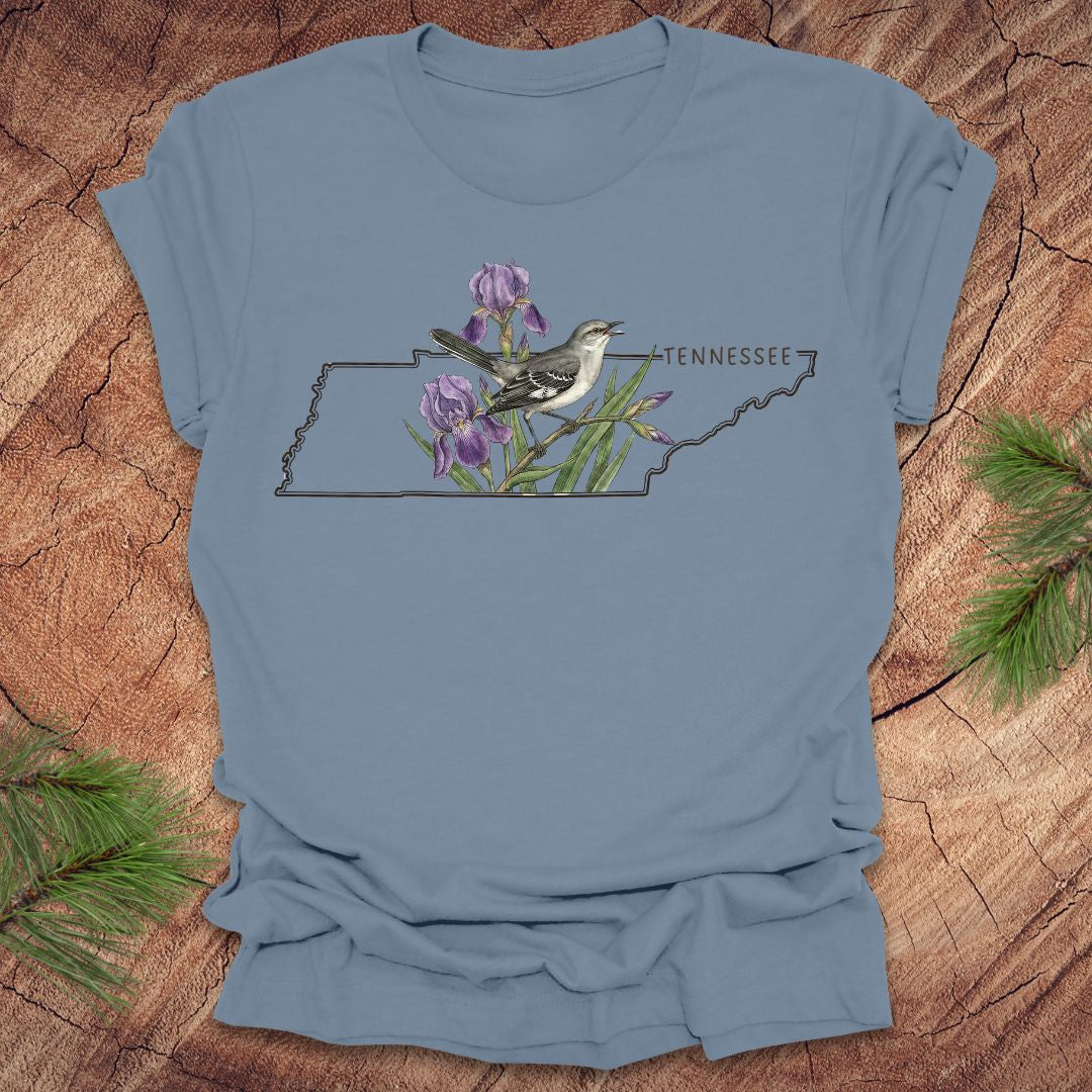 Steel blue t-shirt with a graphic of Tennessee's state shape featuring a mockingbird and iris flowers on a wooden surface.