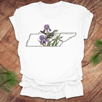 White t-shirt with a graphic of Tennessee's state shape featuring a mockingbird and iris flowers on a wooden surface.