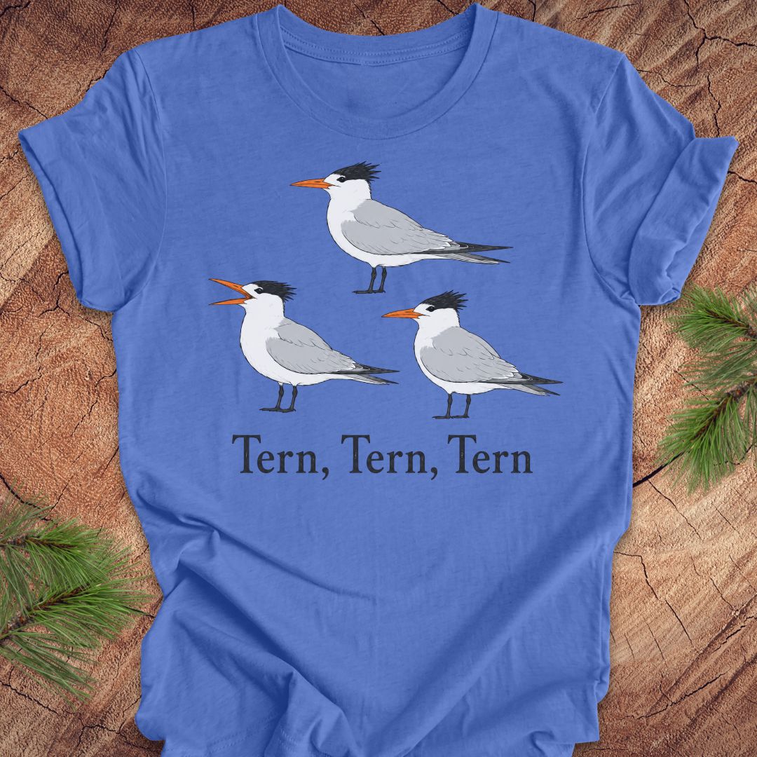 A blue t-shirt with three tern birds and text, 'tern, tern, tern' on a wood background. 
