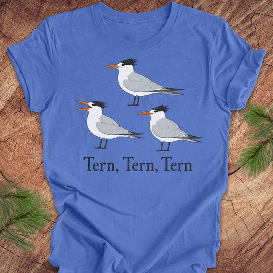 A blue t-shirt with three tern birds and text, 'tern, tern, tern' on a wood background. 