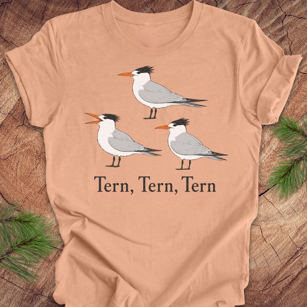 A peach t-shirt with three tern birds and text, 'tern, tern, tern' on a wood background. 