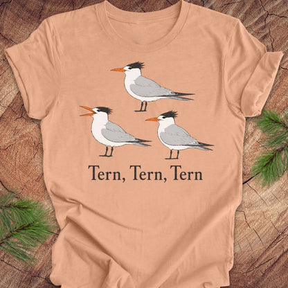 A peach t-shirt with three tern birds and text, 'tern, tern, tern' on a wood background. 