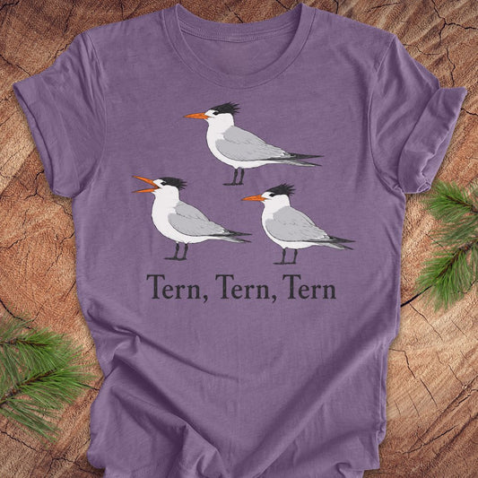 A purple t-shirt with three tern birds and text, 'tern, tern, tern' on a wood background. 