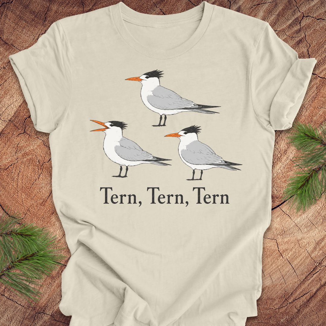 A beige t-shirt with three tern birds and text, 'tern, tern, tern' on a wood background. 