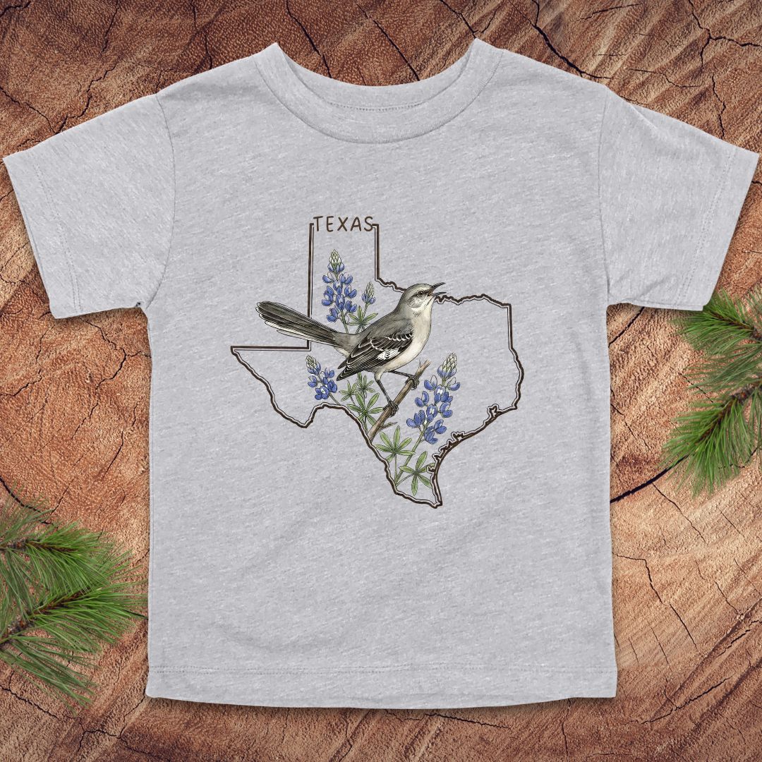 Gray toddler t-shirt with Texas state outline and mockingbird on a wood background.