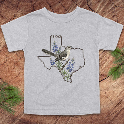Gray toddler t-shirt with Texas state outline and mockingbird on a wood background.