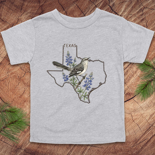 Gray toddler t-shirt with Texas state outline and mockingbird on a wood background.
