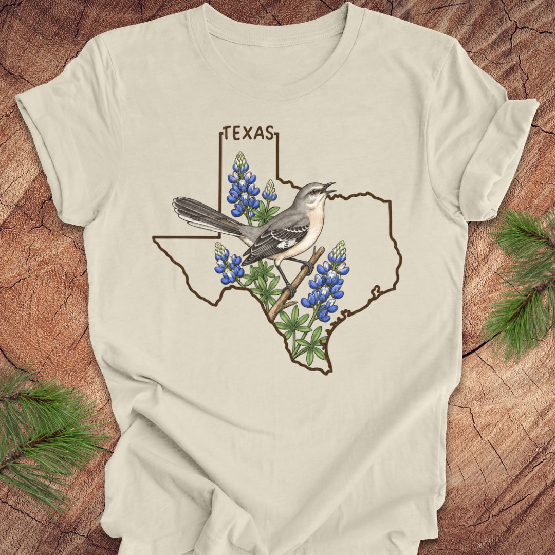 An off-white t-shirt laid flat on a rustic wood background, featuring a graphic of a Texas state outline with a gray bird perched on bluebonnet flowers and the word 'TEXAS' above.