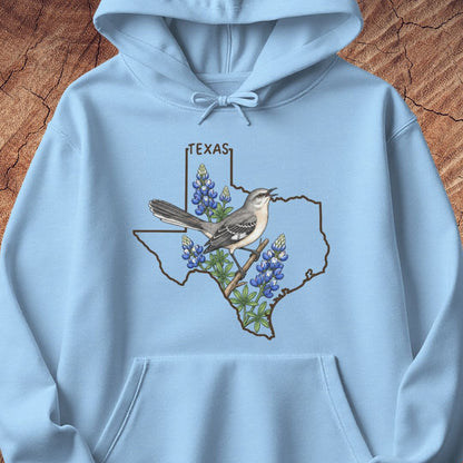 A light blue pullover hoodie laid flat on a textured brown wood background. The front features a graphic of the state outline of Texas with a mockingbird perched on a branch among bluebonnets, and the word 'TEXAS' above.