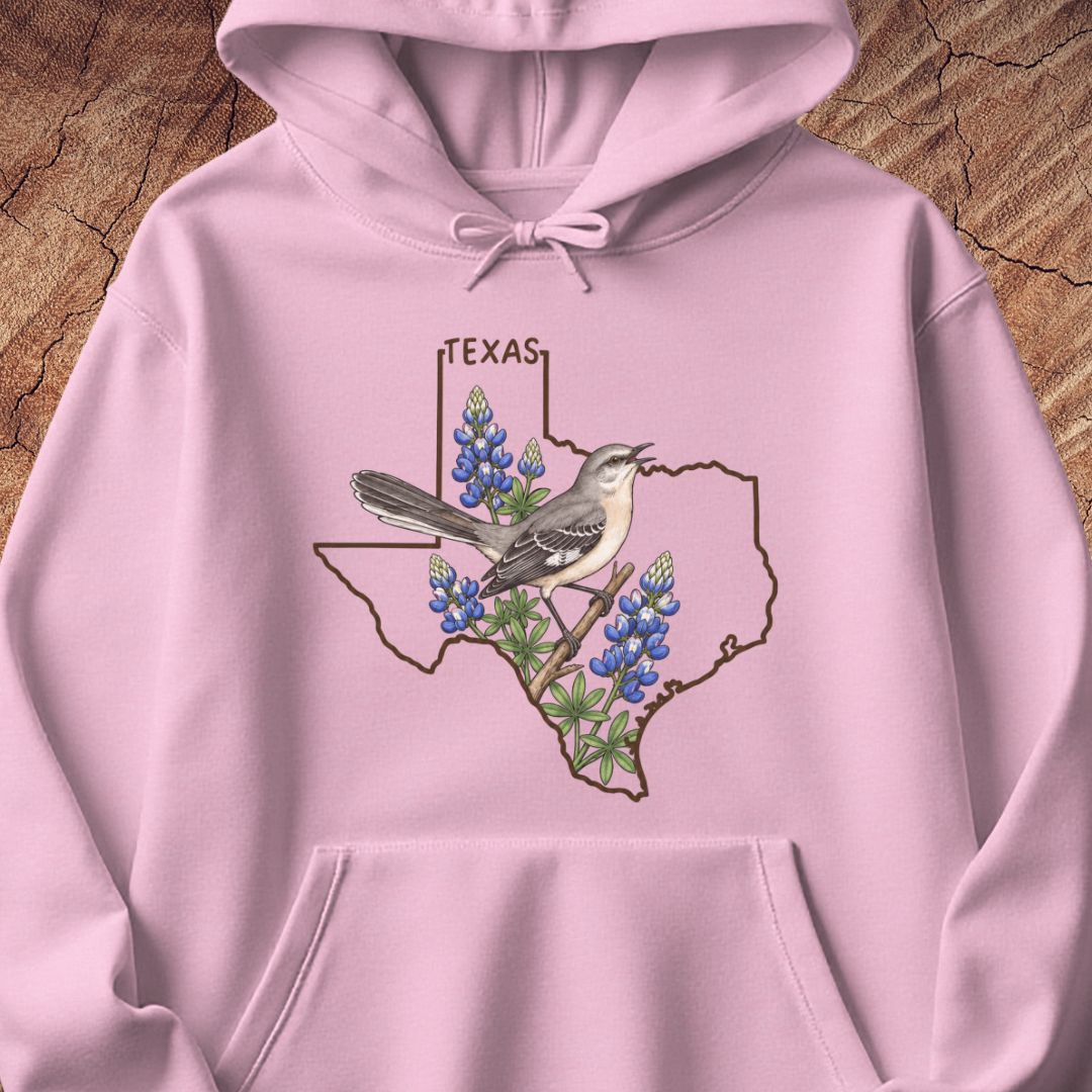 A light pink pullover hoodie laid flat on a textured wood background, featuring a graphic of a gray and white bird perched on bluebonnets inside the outline of Texas, with the word TEXAS above.