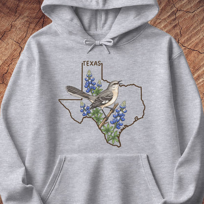 A light heather gray pullover hoodie featuring a graphic print of the state outline of Texas with bluebonnets, a gray-and-white bird, and the word 'TEXAS' above it, shown against a rustic wood background.