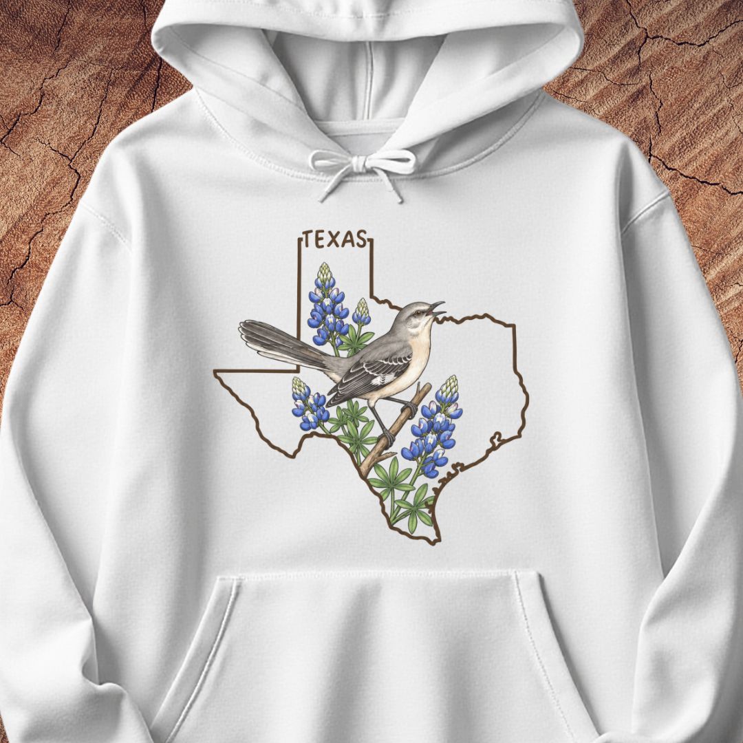 A white pullover hoodie with a front graphic showing the outline of Texas filled with bluebonnets and a bird, displayed on a textured brown wood background.