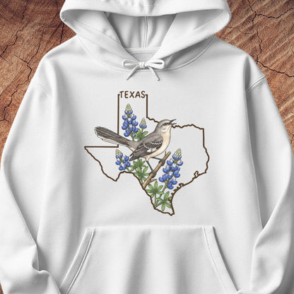 A white pullover hoodie with a front graphic showing the outline of Texas filled with bluebonnets and a bird, displayed on a textured brown wood background.