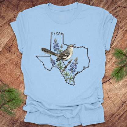 Baby blue t-shirt with Texas state outline with state bird, mockingbird and state flower bluebonnet design on a wooden surface