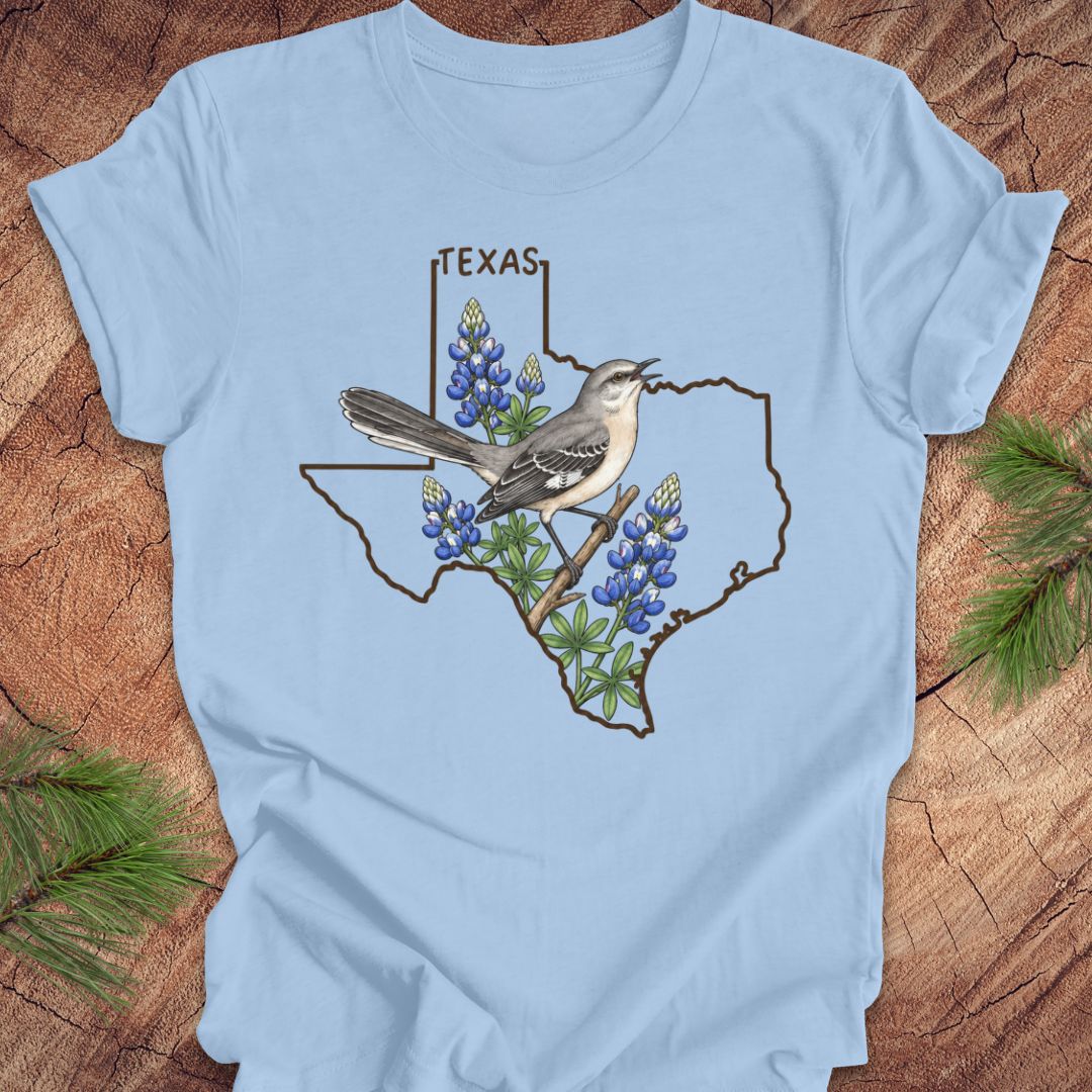 A light blue short-sleeve t-shirt laid flat on a rustic wood surface, featuring a graphic of a Texas state outline with a mockingbird perched on bluebonnets and the word TEXAS above.