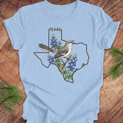 A light blue short-sleeve t-shirt laid flat on a rustic wood surface, featuring a graphic of a Texas state outline with a mockingbird perched on bluebonnets and the word TEXAS above.