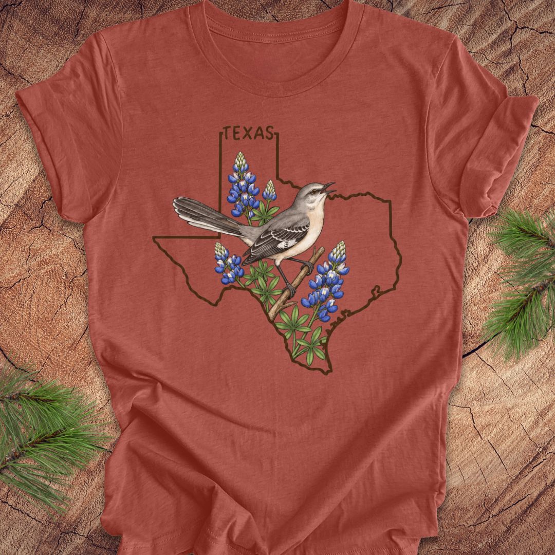 A rust-colored short-sleeve t-shirt laid flat on a wood surface, featuring a graphic of the state outline of Texas with bluebonnets and a gray-and-white bird perched on it. Pine branches are placed around the shirt as styling props.